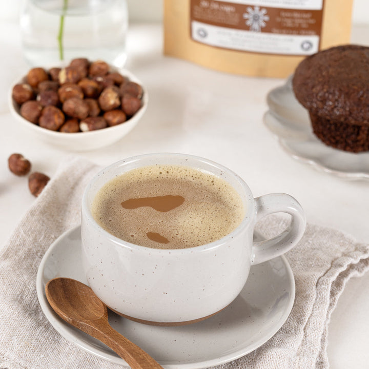Cup of coffee with a wooden spoon on a white saucer, surrounded by coffee beans and a muffin.