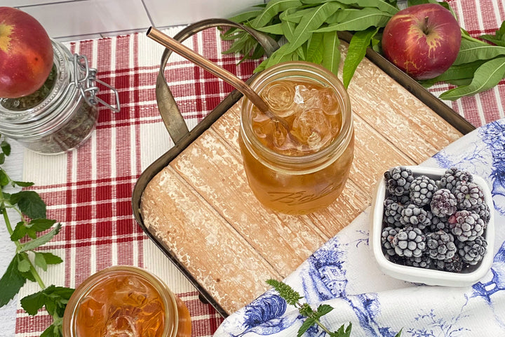 Jars of iced tea, apples, and blackberries on a red checkered tablecloth with a ameriacana chicken cloth and fresh mint.