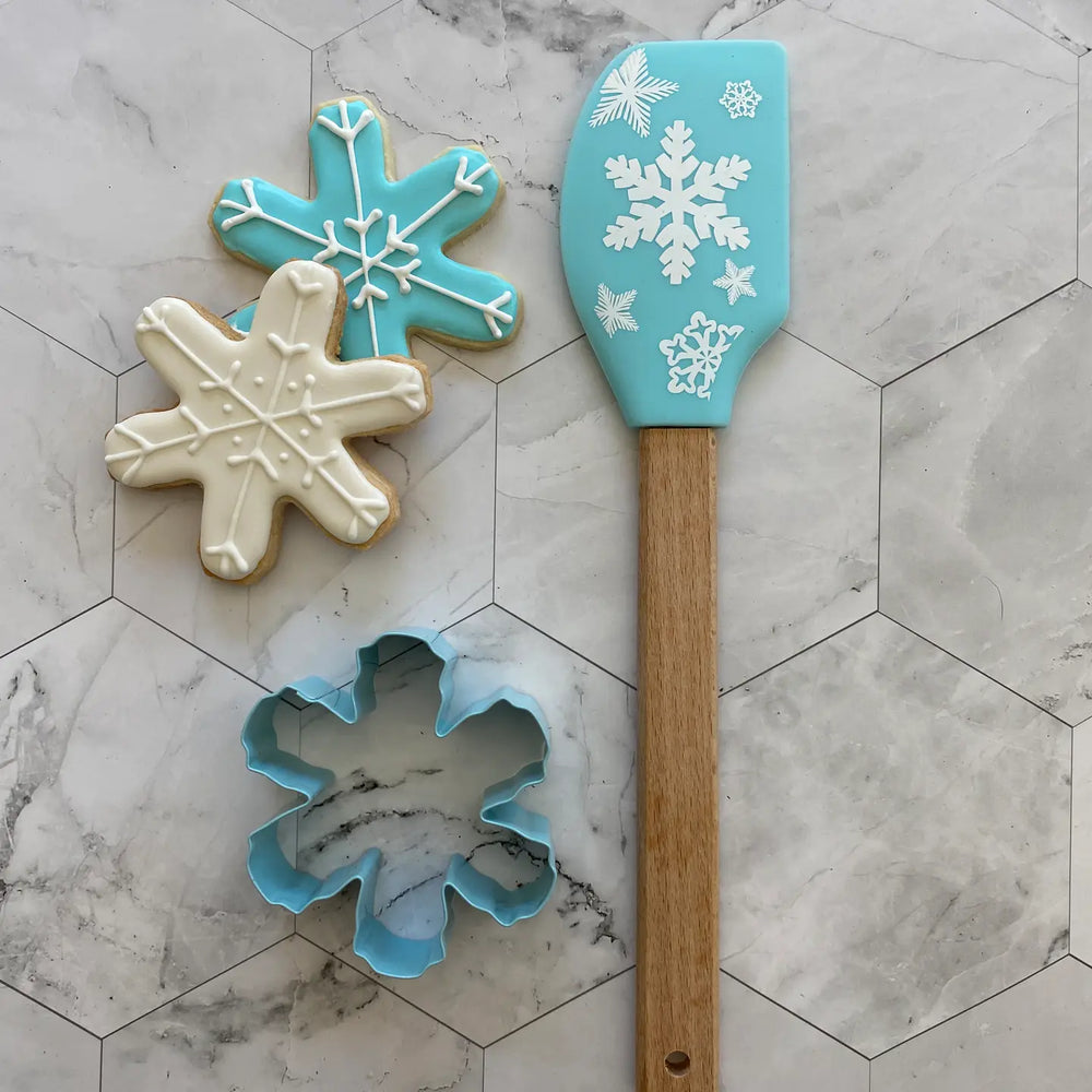Snowflake-shaped cookies, cookie cutter, and spatula with snowflake design on a marble surface