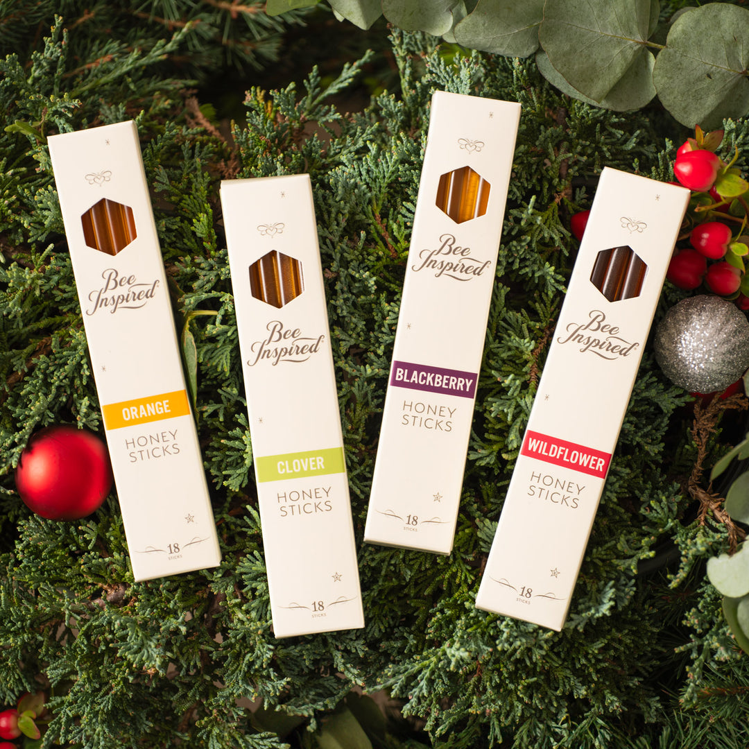 Four packages of 'Bee Inspired' honey sticks on a green leafy background with Christmas decorations.